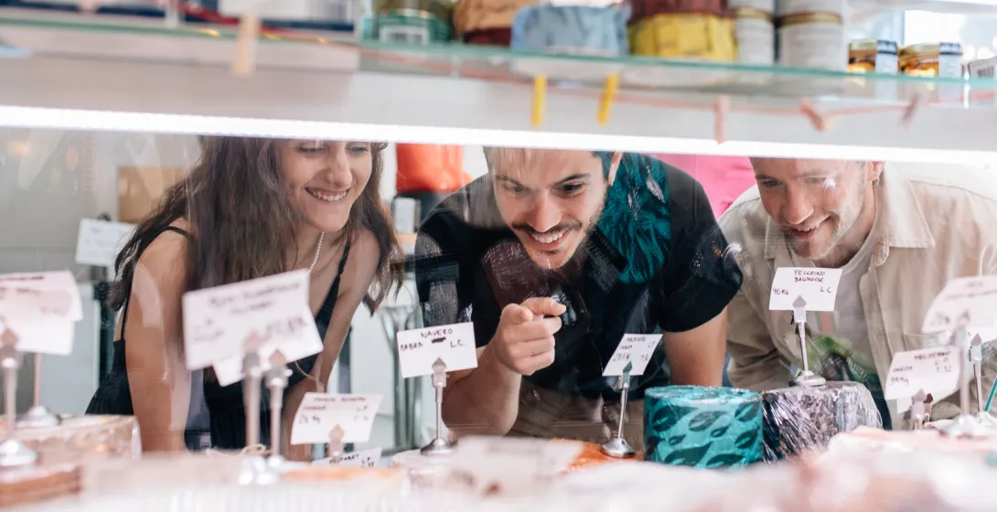 Tres jóvenes sonríen mientras miran una selección de quesos a través de un mostrador de cristal en una tienda.