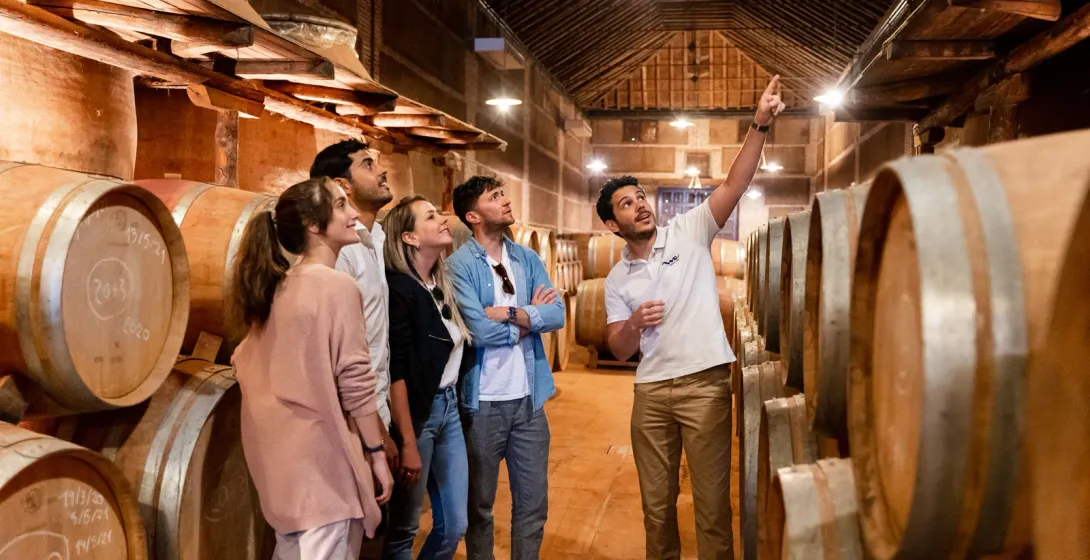 Un guía turístico explica algo a un grupo de visitantes en una bodega rodeada de grandes barricas de madera.