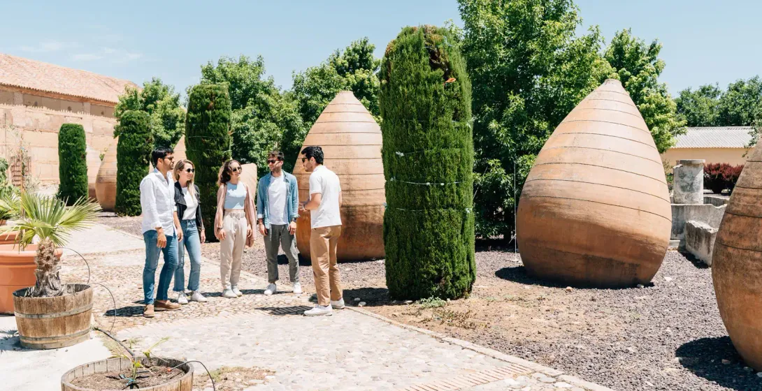 Un guía turístico habla con un pequeño grupo de personas en un patio soleado con grandes tinajas de barro de estilo antiguo.