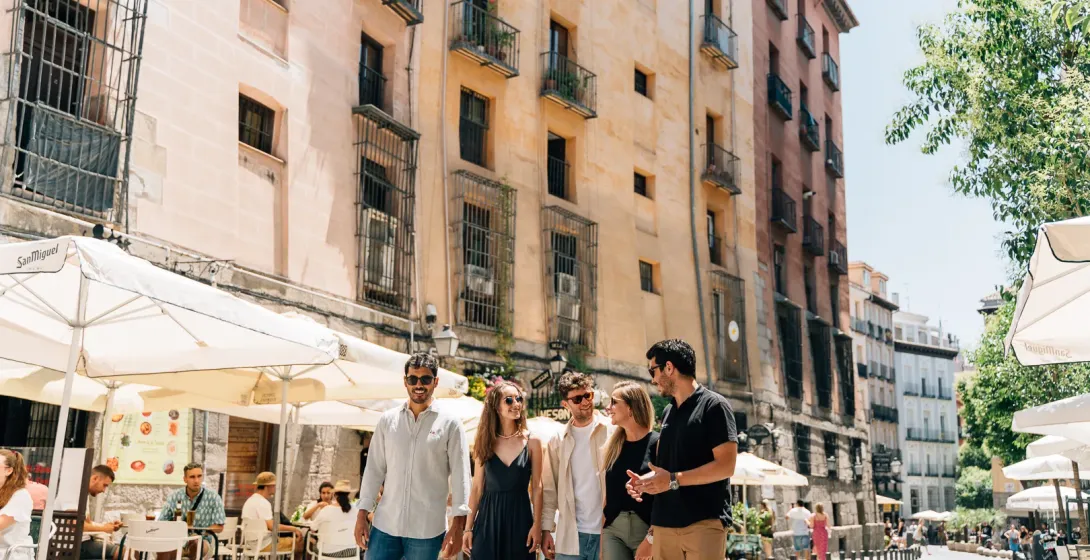 Un grupo de cinco jóvenes camina por una soleada calle de Madrid bordeada de mesas de café y edificios históricos.