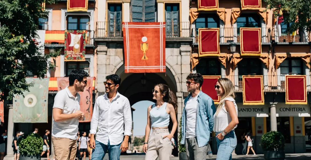 Un grupo de cinco jóvenes en una plaza soleada, frente a un edificio decorado con estandartes rojos y dorados.