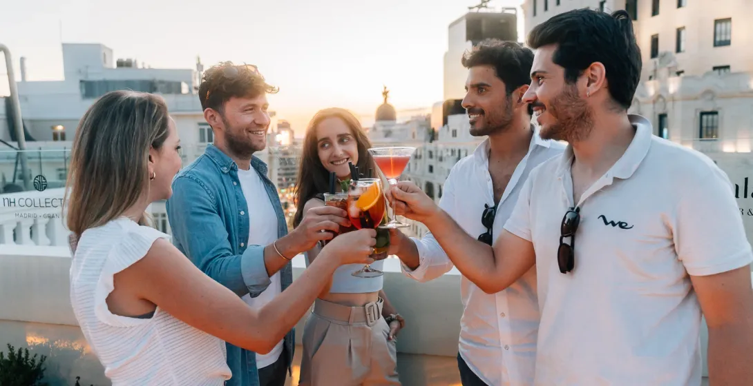 Cinco jóvenes amigos brindan con cócteles de colores en una azotea de Madrid al atardecer.