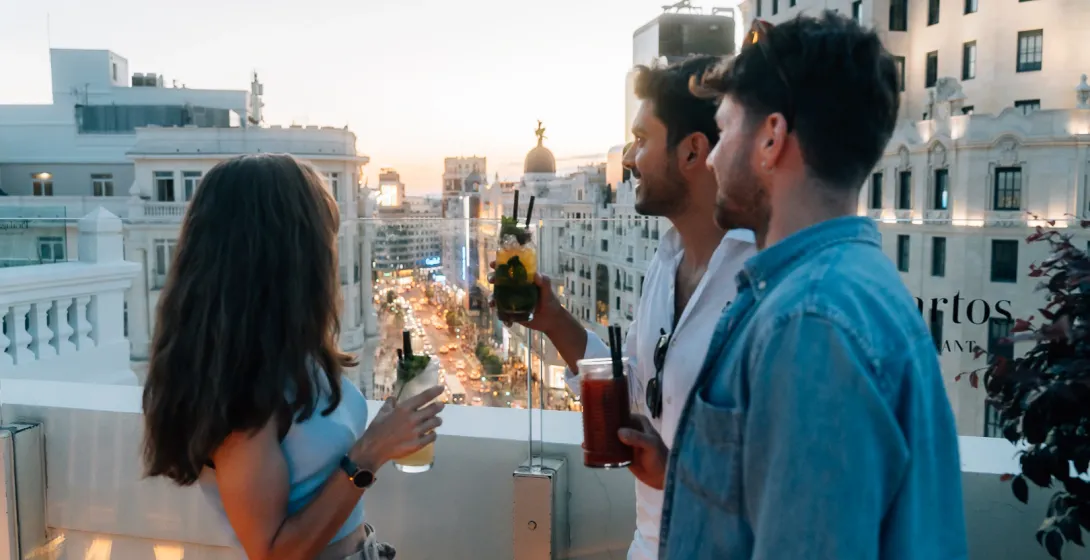 Tres amigos con cócteles en una azotea con vistas a las concurridas calles de Madrid al atardecer.