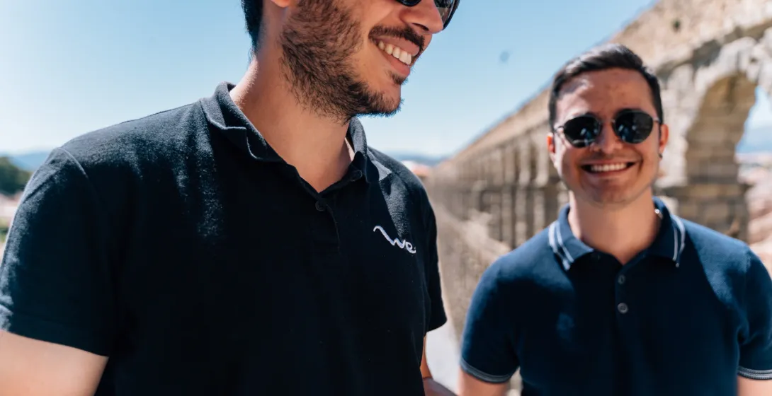 Dos jóvenes con gafas de sol sonríen frente al Acueducto de Segovia en un día soleado.