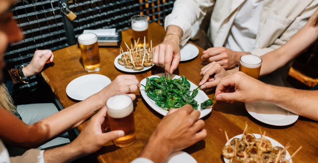 Amigos compartiendo tapas, incluyendo pimientos de Padrón, y bebiendo cerveza en una mesa de madera en un bar.
