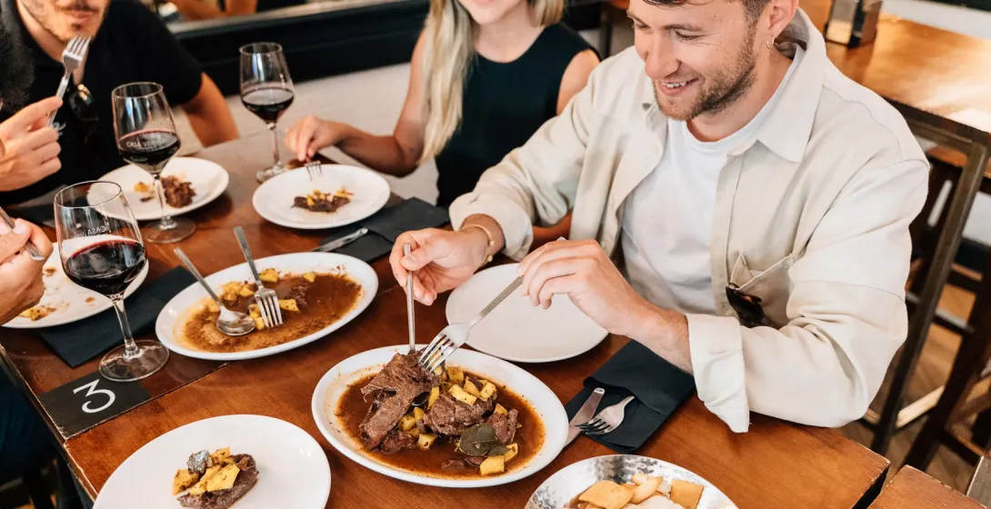 Tres personas en una mesa de madera compartiendo platos de estofado de ternera y bebiendo vino tinto en un restaurante.