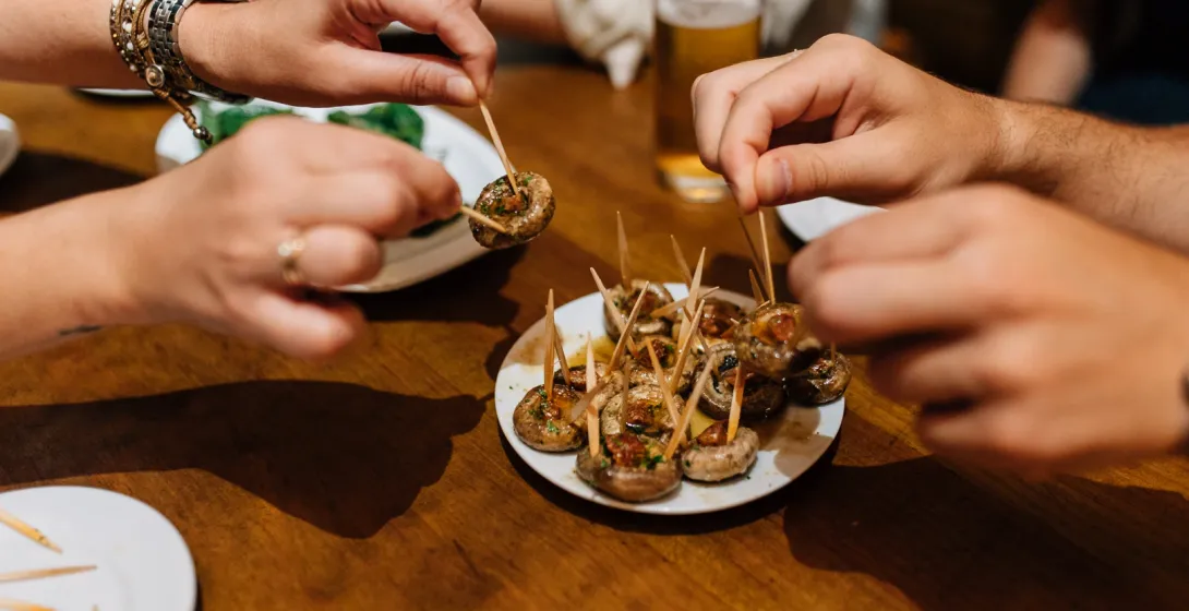 Manos cogiendo tapas de champiñones con palillos de un plato en una mesa de madera.