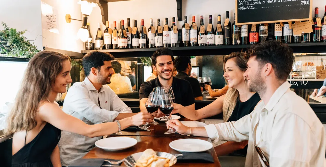 Un grupo de cinco amigos sentados a una mesa en un bar, sonriendo y brindando con copas de vino tinto.