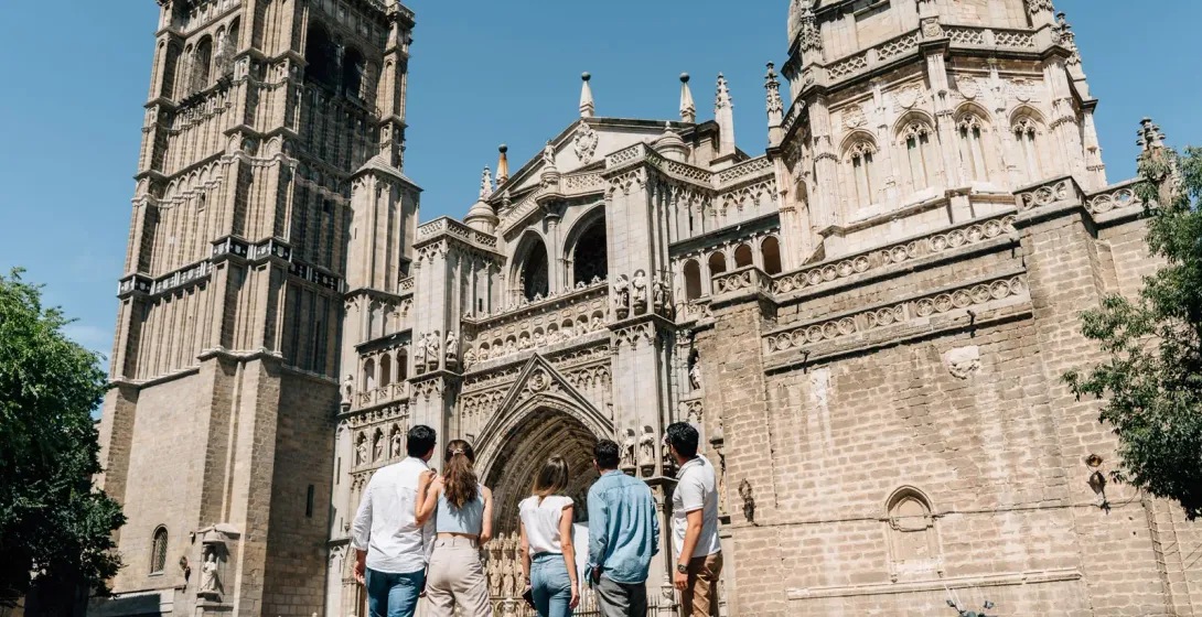 Un grupo de cinco personas, vistas desde atrás, admiran la intrincada fachada de piedra de la Catedral de Toledo en un día...