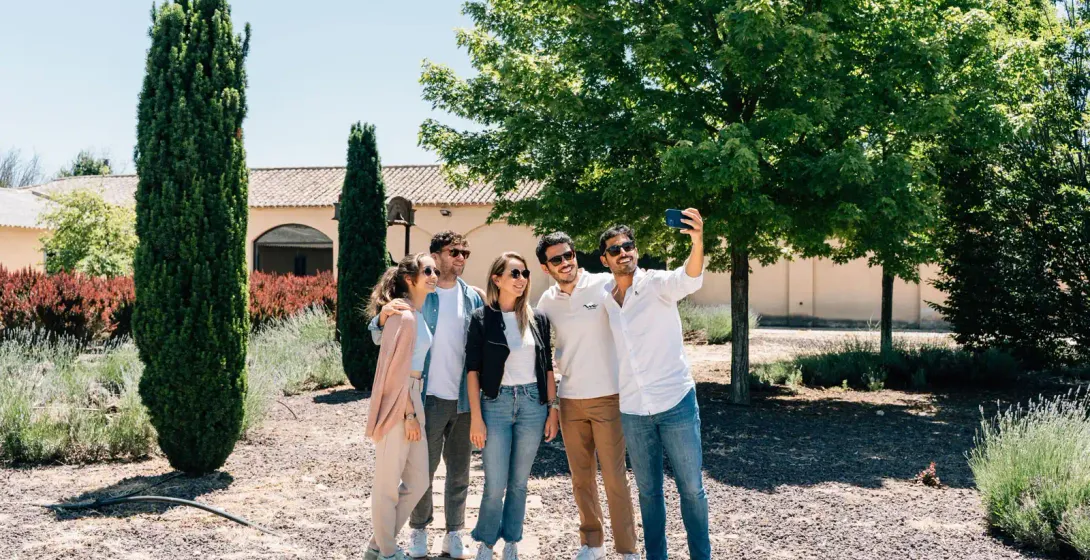 Un grupo de cinco amigos se hace un selfi sonriente en un jardín soleado con cipreses y una villa al fondo.