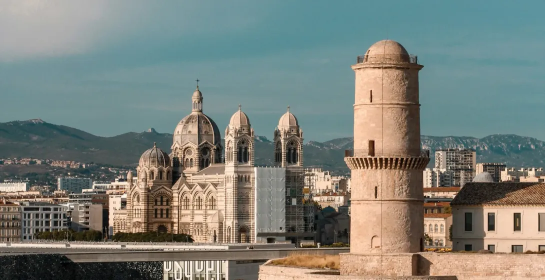 Marseille's Fort Saint-Jean tower and walls by the sea, with Cathédrale La Major, Mucem museum, and distant mountains.