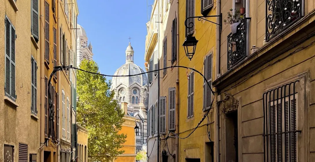 Narrow street in Marseille lined with tall yellow buildings with shutters, leading towards the dome of Notre-Dame de la Ga...