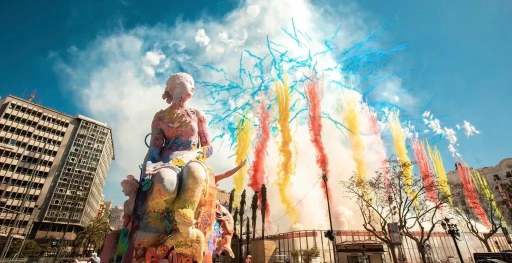 Una gran y colorida estatua de una 'falla' con un espectáculo de fuegos artificiales diurnos de humo de colores detrás de ...