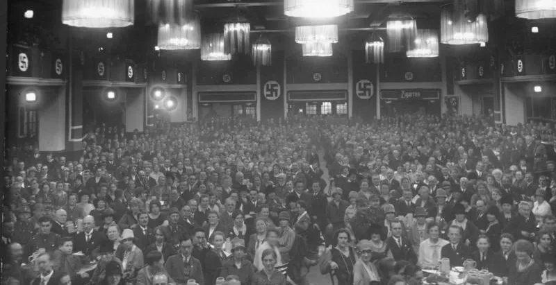 Foto en blanco y negro de un gran salón abarrotado de gente sentada en mesas. Banderas con esvásticas y letrero de 'Zigare...