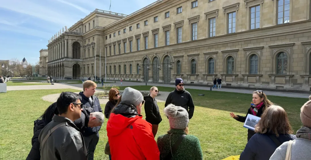 A tour guide with a yellow Walkative umbrella speaks to a group on a lawn in front of a large neoclassical building in Mun...