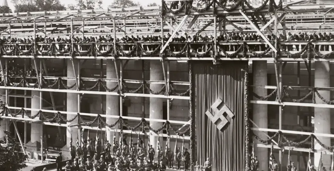 Schwarz-weiß-Foto eines im Bau befindlichen Gebäudes, geschmückt mit Girlanden und einem großen Hakenkreuzbanner. Menschen...