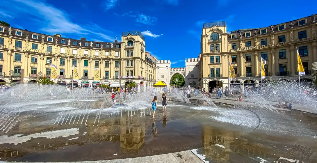 People enjoying the large fountain at Karlsplatz in Munich, with Karlstor archway and historic buildings in the background...