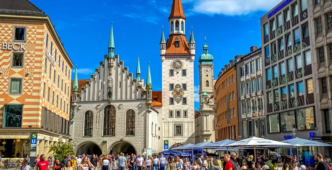 Crowded Marienplatz in Munich under a blue sky, featuring the New Town Hall with its tall spire and the Old Town Hall.