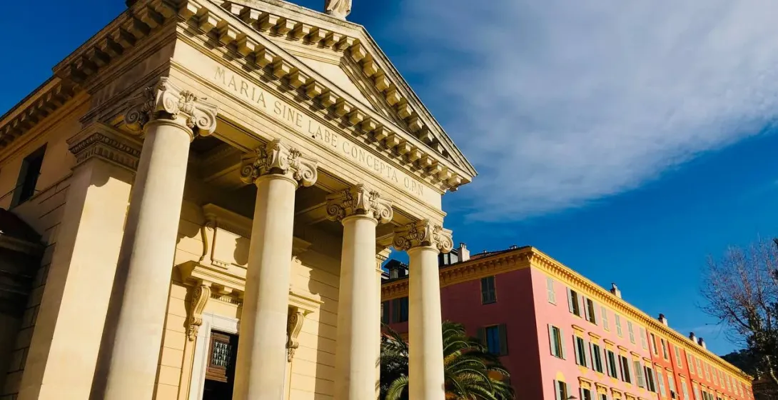 Neo-classical chapel with columns, pediment, and statue under a blue sky, next to a pink building with arcades in Nice.