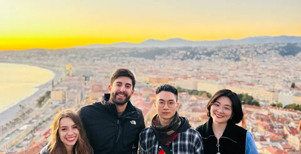 Four people posing on a viewpoint overlooking Nice city, the bay, and mountains at sunset. Golden light illuminates the ho...