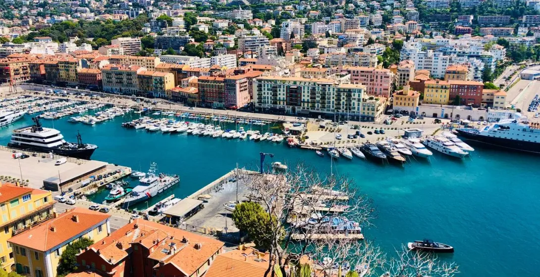 Panoramic view of Nice's Port Lympia with numerous yachts, colorful waterfront buildings, and green hills under a clear bl...