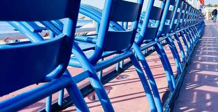 A row of iconic blue chairs on the Promenade des Anglais in Nice, facing the sea under a clear blue sky. Shadows extend on...