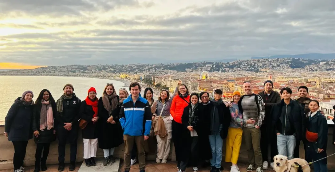 Diverse group and a golden retriever dog on a tiled terrace overlooking Nice city, bay, and coastline at sunset.