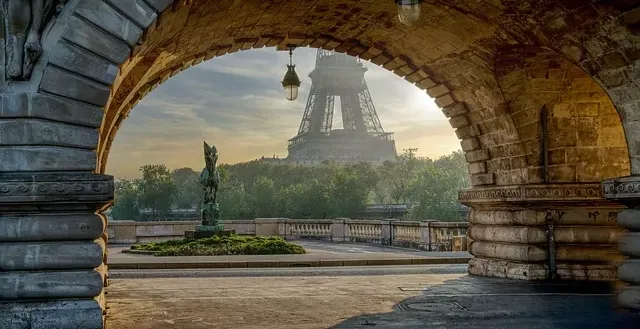 Eiffel Tower seen through a stone archway of a bridge, with a green statue and trees in the foreground under a bright sky.