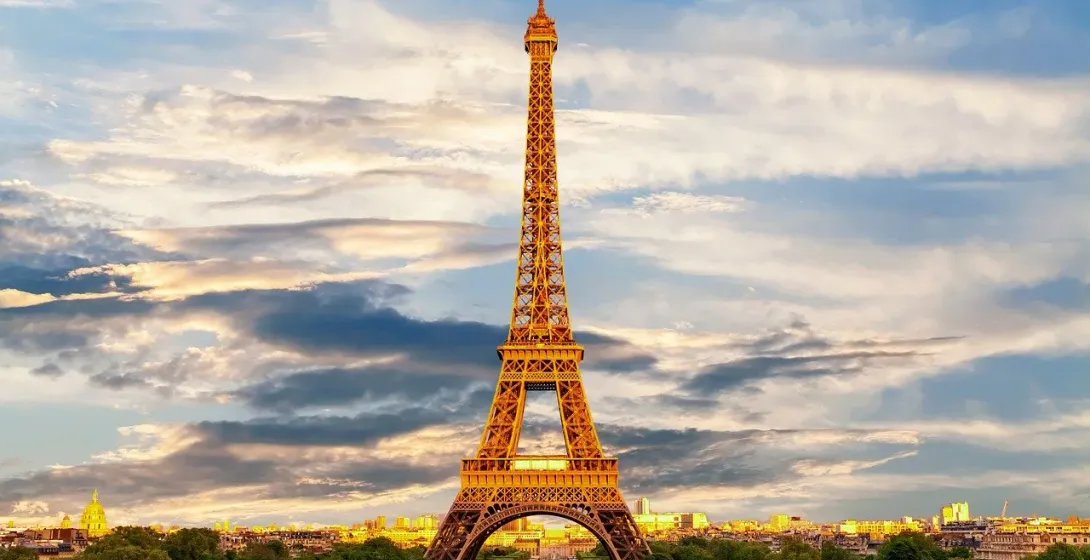 The Eiffel Tower rises above Paris at golden hour, with city buildings, green trees, and scattered clouds in the sky.