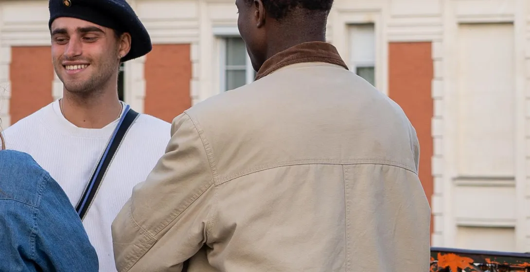 Two people, one smiling in a beret next to another from behind, with love locks and a building in background.