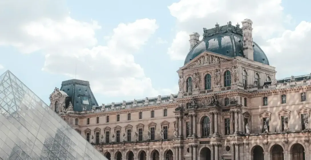 The Louvre Museum palace building with a dark dome and the glass pyramid in the foreground under a cloudy sky.