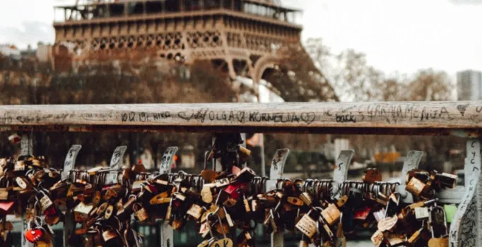 Numerous 'love locks' of various sizes and colors are attached to a grey metal bridge railing. The Eiffel Tower is blurred...