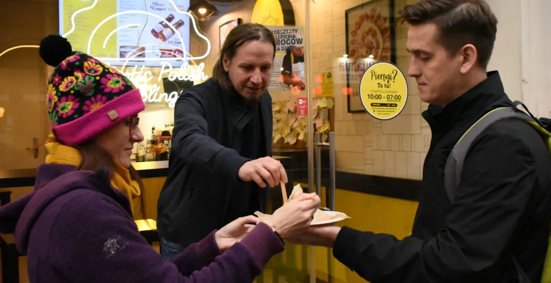 Three people standing outside a dumpling shop in Krakow, sharing pierogi from a paper plate.