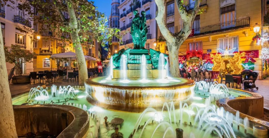 Una fuente iluminada en la Plaza Rodrigo Botet de Valencia, rodeada de árboles y edificios con luces de Navidad.