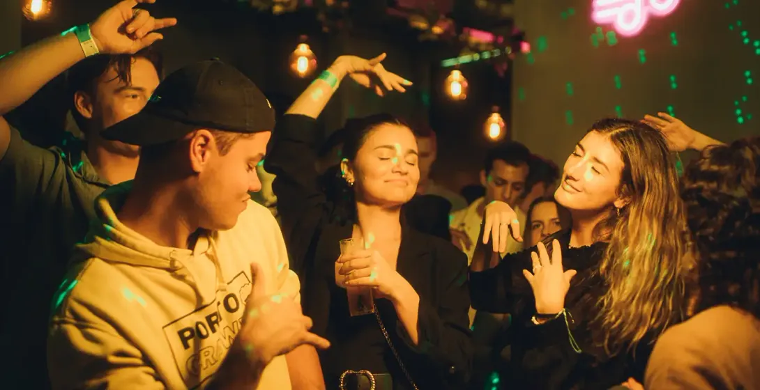 Menschen tanzen in einem schwach beleuchteten Club oder einer Bar, mit grünen Laserlichtern und einem pinken Neon-Musiknot...