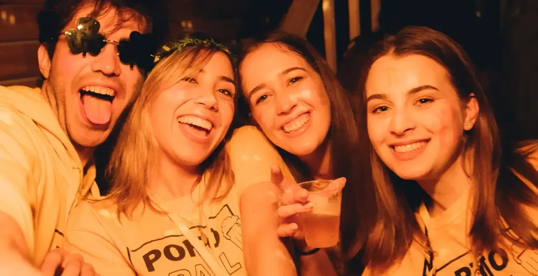 Vier junge Leute, zwei Männer und zwei Frauen, lächeln in einer Bar. Sie tragen gelbe 'PORTO PUB CRAWL' T-Shirts und halte...