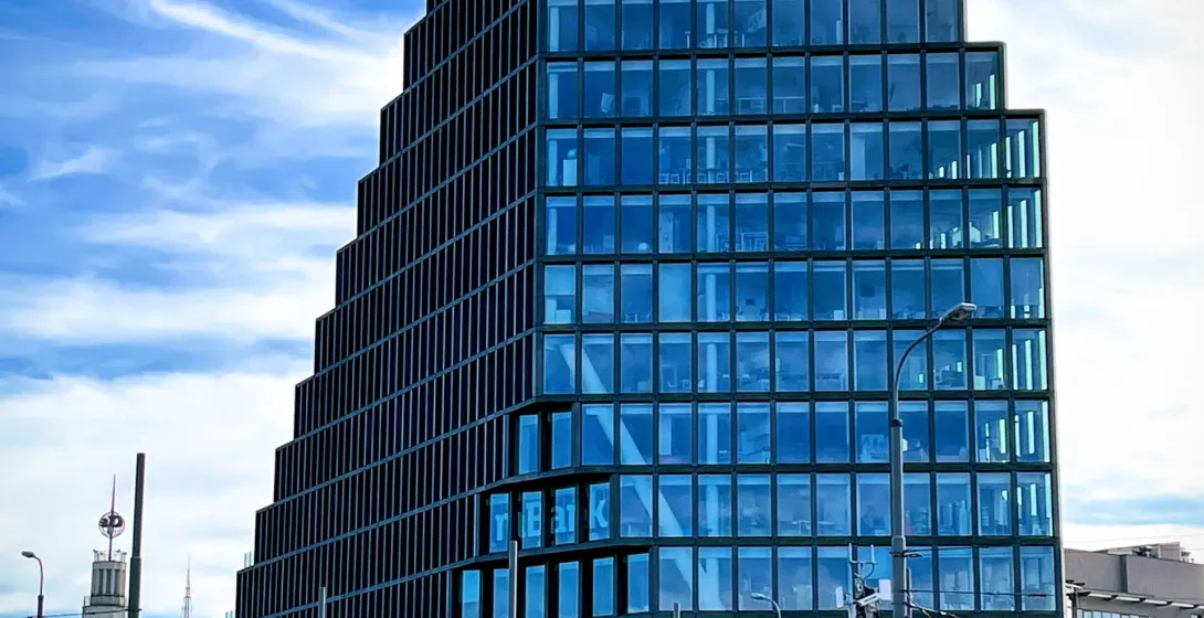 Moderno edificio Bałtyk en Poznań con fachada de vidrio escalonada, bajo cielo azul, con calle y torre histórica visibles.