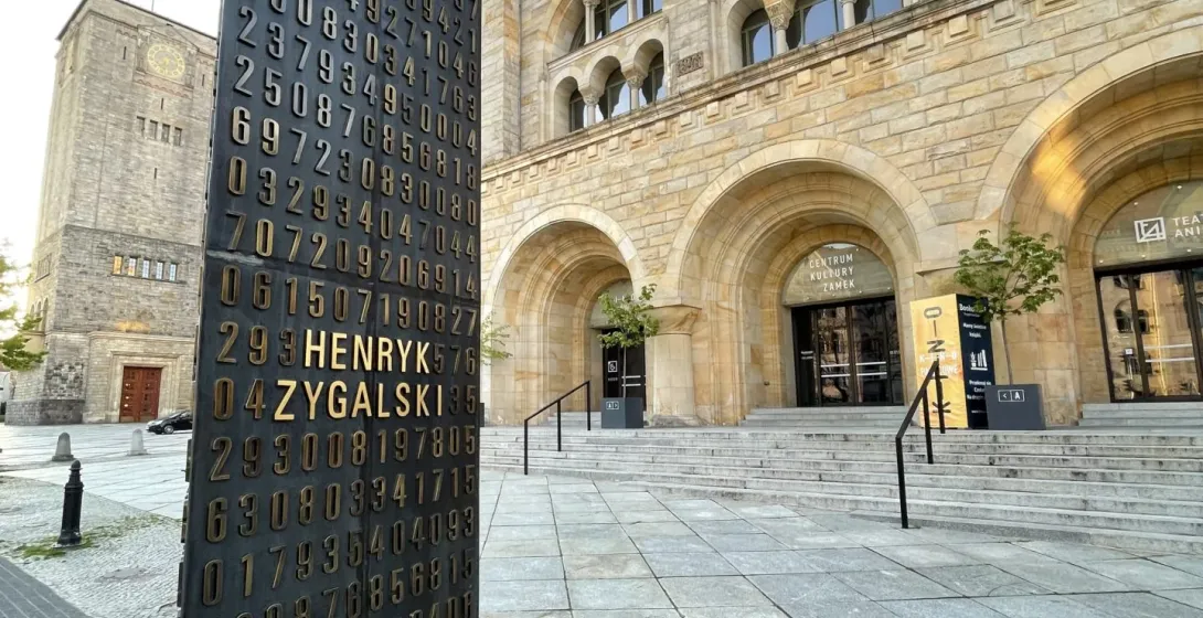 Dunkles Denkmal mit goldenen Zahlen und 'HENRYK ZYGALSKI' auf einem gepflasterten Platz, vor dem Posener Kaiserschloss mit...