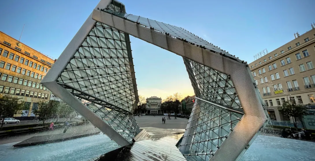 Fuente moderna de vidrio y metal con chorros de agua, en forma de cubo abierto, en una plaza de la ciudad al anochecer. Ed...