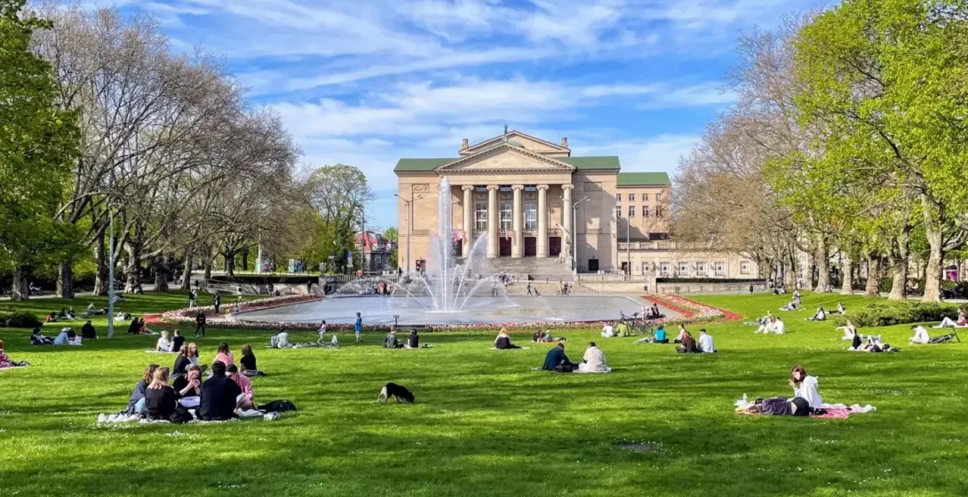 Grand Theatre building, fountain, and people relaxing on green grass in Poznań park on a sunny day.
