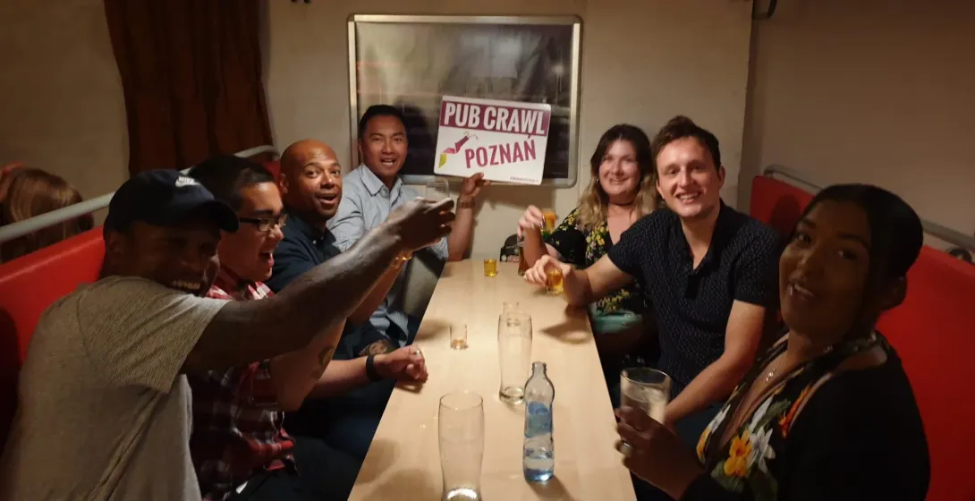Diverse group of young adults smiling at a table in a pub, some holding drinks. One person displays a 'Pub Crawl Poznan' s...