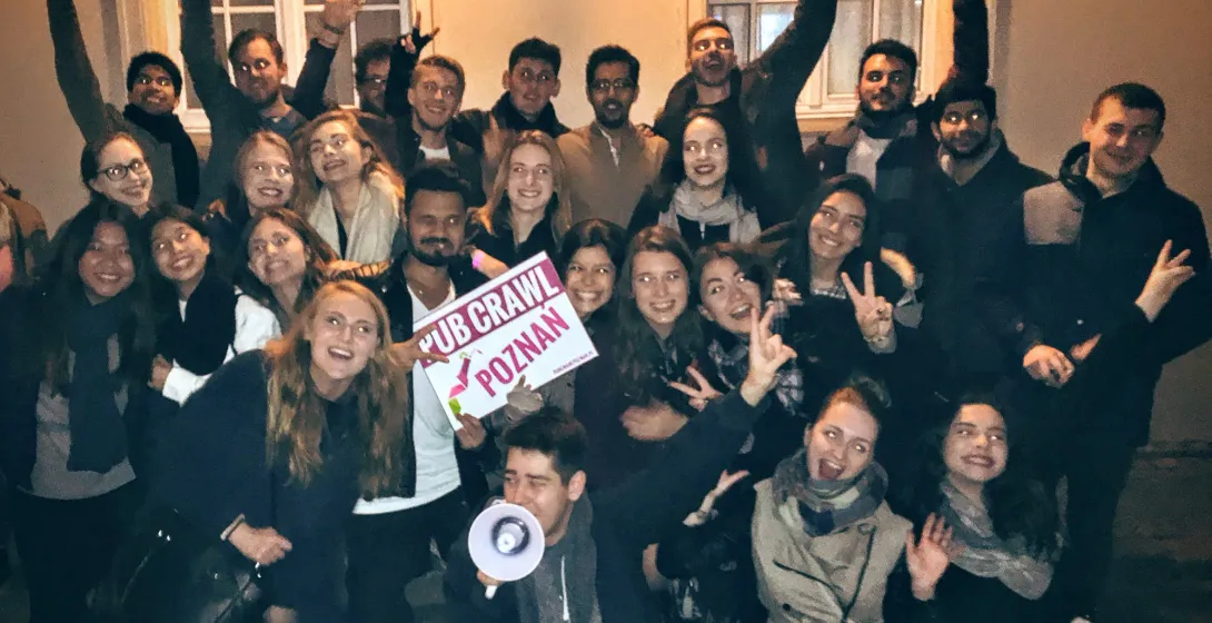 A group of young adults smiling and waving on a pub crawl, holding a "PUB CRAWL POZNAN" sign. One person holds a megaphone.