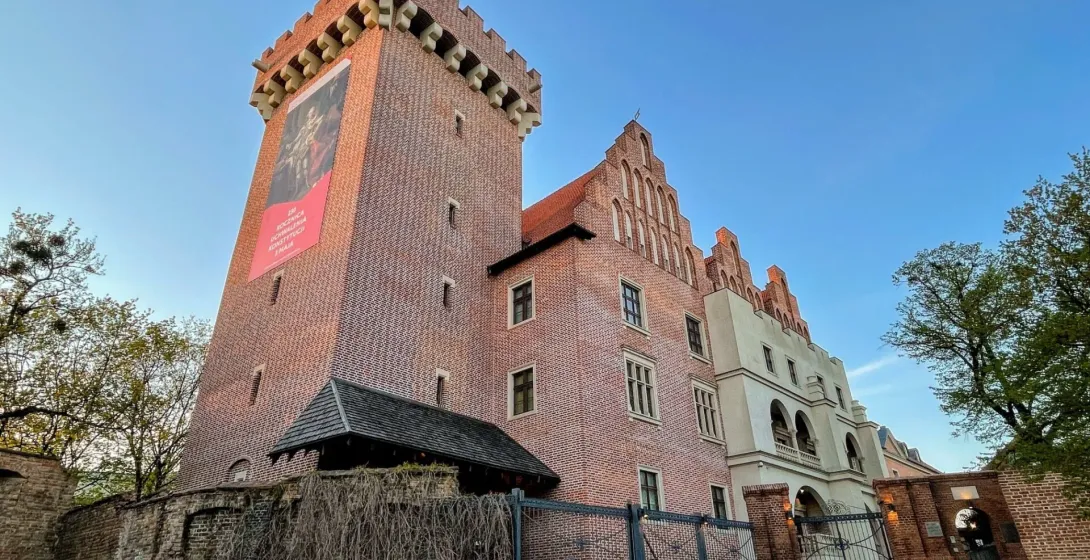 Widok na Zamek Królewski w Poznaniu, ceglany budynek z wysoką wieżą, szczytową fasadą i starym murem, pod błękitnym niebem.