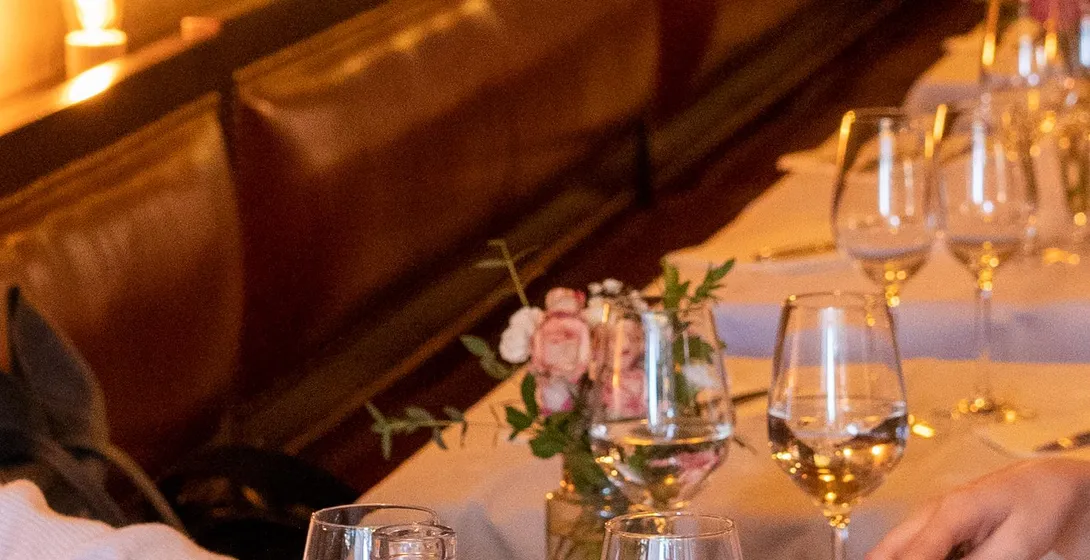 Close-up of people dining at a well-lit European restaurant, with bread, water, wine glasses, and cheese on table.