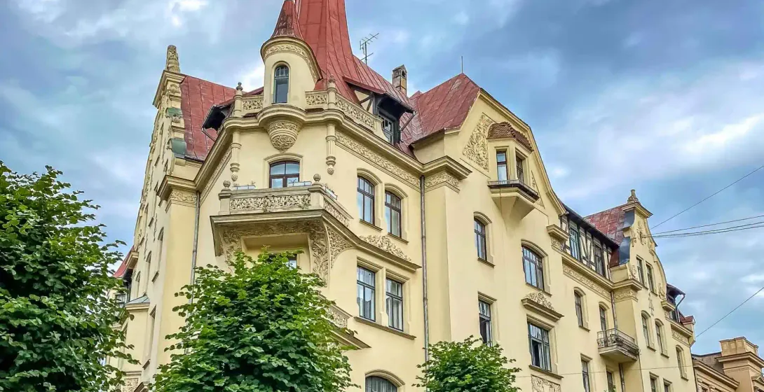 Edificio Art Nouveau ornamentado en Riga con fachada amarilla, techo rojo y torre, enmarcado por árboles verdes bajo cielo...