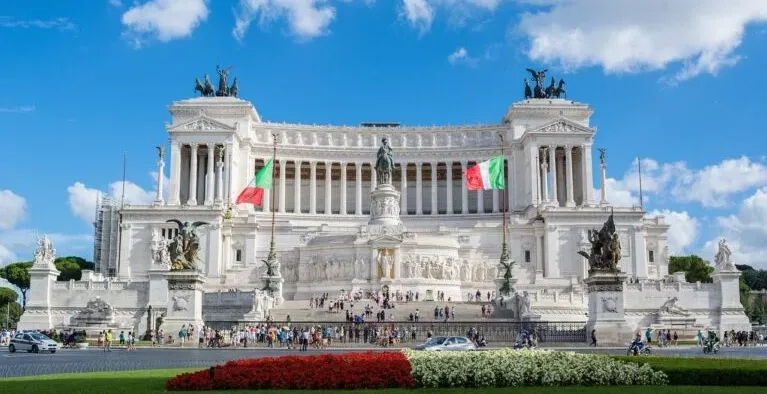 Gran Altar de la Patria de mármol blanco en Roma bajo un cielo azul con nubes. Banderas italianas a cada lado. Gente en la...