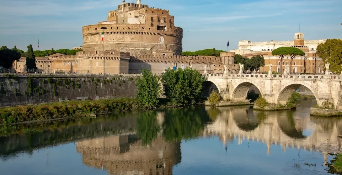 Zamek Świętego Anioła i Most Świętego Anioła nad Tybrem, z odbiciami w wodzie pod błękitnym niebem.