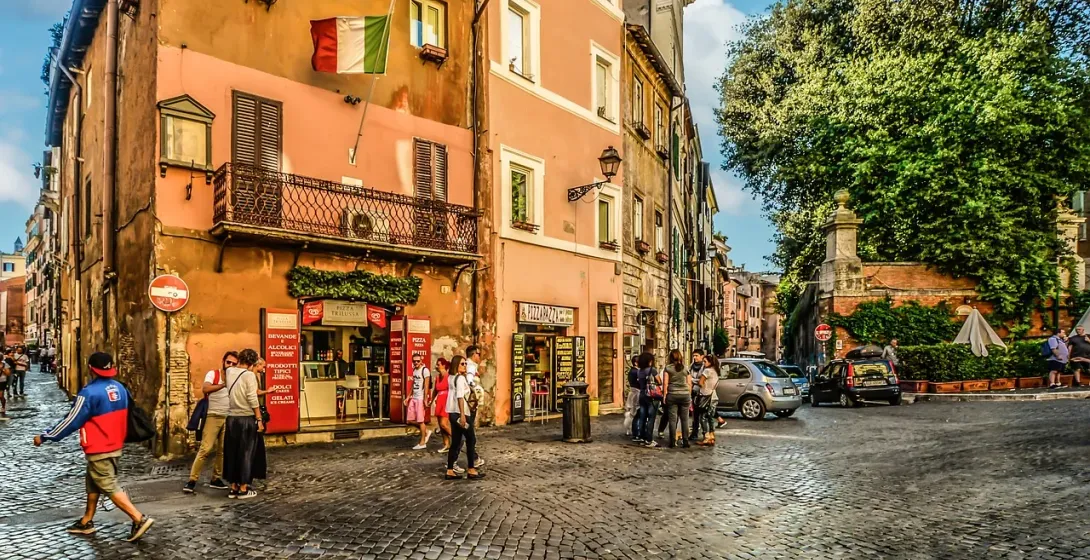Calle adoquinada en Roma con edificios históricos, bandera italiana, personas caminando y coches aparcados bajo un cielo a...
