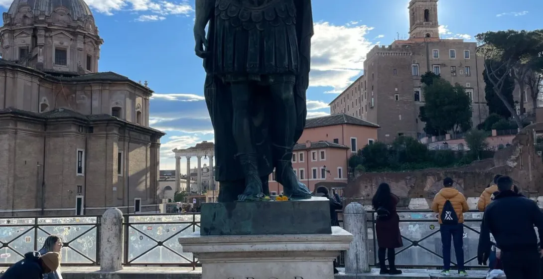 Bronzestatue von Julius Cäsar in Rom, mit Ruinen des Forum Romanum und einer Kuppelkirche unter blauem Himmel.