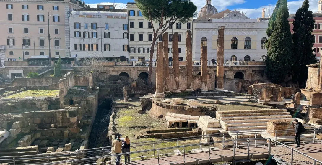 Widok starożytnych rzymskich ruin z odkrytymi poziomami, kamiennymi kolumnami i nowoczesną drewnianą kładką. Budynki miejs...